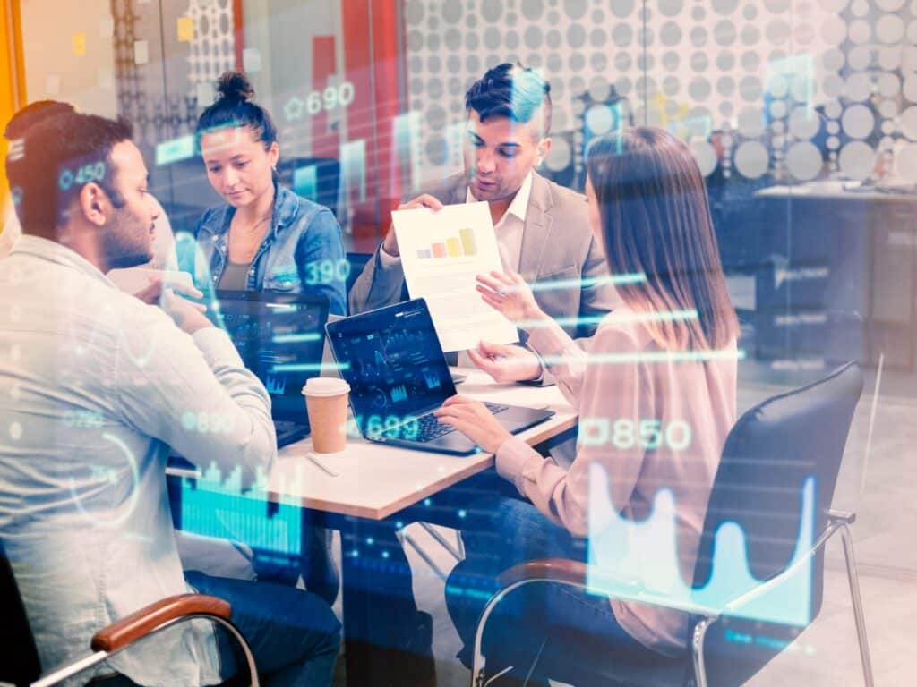 Four professionals in a modern office sit around a table with laptops and a printed report showing a bar chart, while a glass wall behind them displays translucent financial data overlays.